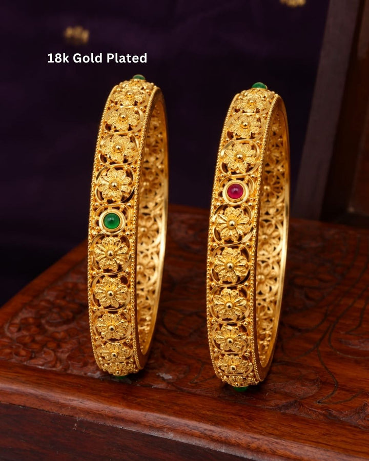 Elegant 18K gold plated floral bangles displayed on a carved wooden jewellery box, showcasing ornate gold craftsmanship and green and ruby stone details.