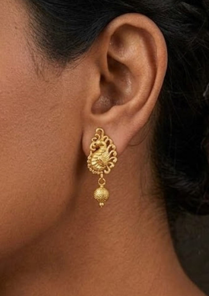 Close up of south indian women ear wearing a Gold plated stud earrings featuring a traditional upright peacock design. The body of the peacock is crafted with clean, textured gold details, and a small, round textured gold ball hangs gently from the bottom of the bird shape, providing a simple, classic look suitable for daily wear, festivals, and family functions.