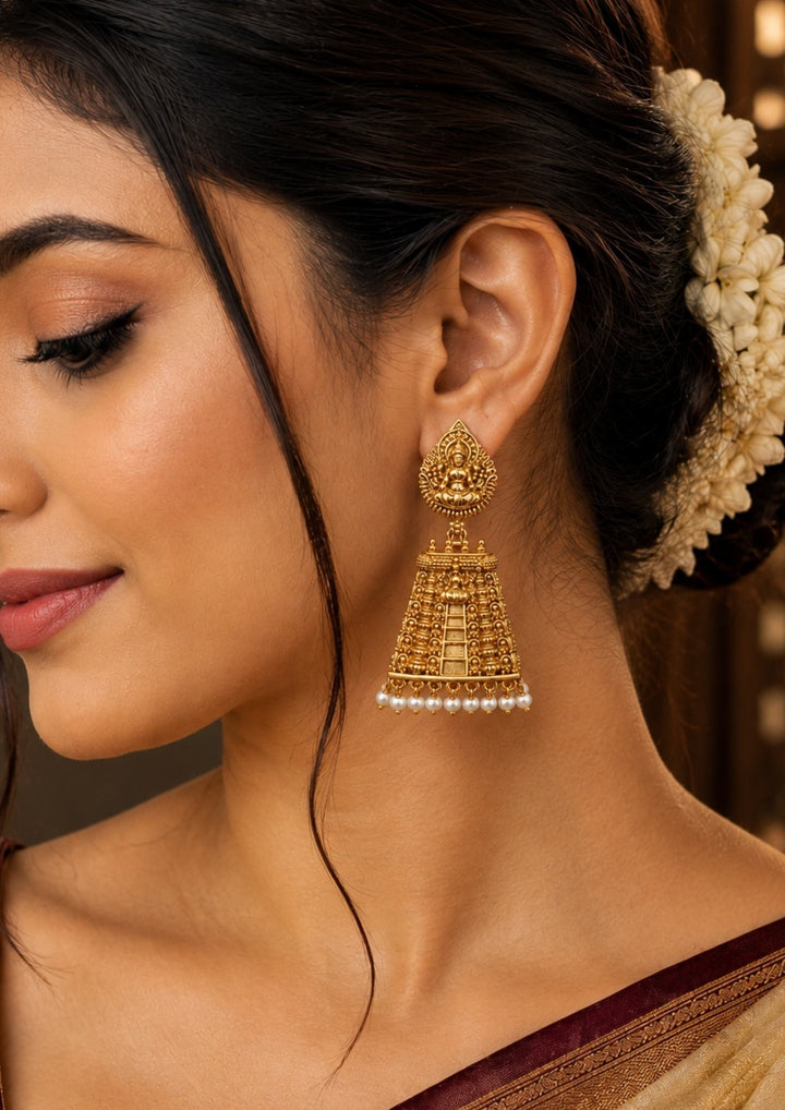 South Indian woman wearing Laxmi Jhumka Gold Plated temple earrings with Goddess Laxmi design, antique gold finish, and pearl hanging jhumka pattern that gives a grand traditional jewelry look. The earrings designed with temple style carvings with dome shaped jhumka detailing and elegant pearl drops that match beautifully with sarees and ethnic outfits. These lightweight earrings come with sweat proof finish, anti-fade colour, skin-friendly quality, and comfortable fitting suitable for weddings