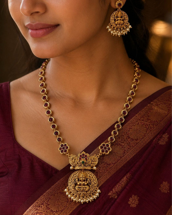 Gold-toned traditional Indian jewelry set featuring a necklace and matching earring with green and red stones, worn by a model in a beige saree.