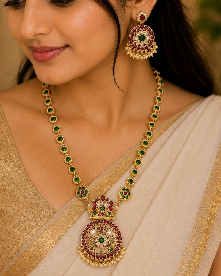 Gold-toned traditional Indian jewelry set featuring a necklace and matching earring with green and red stones, worn by a model in a beige saree.