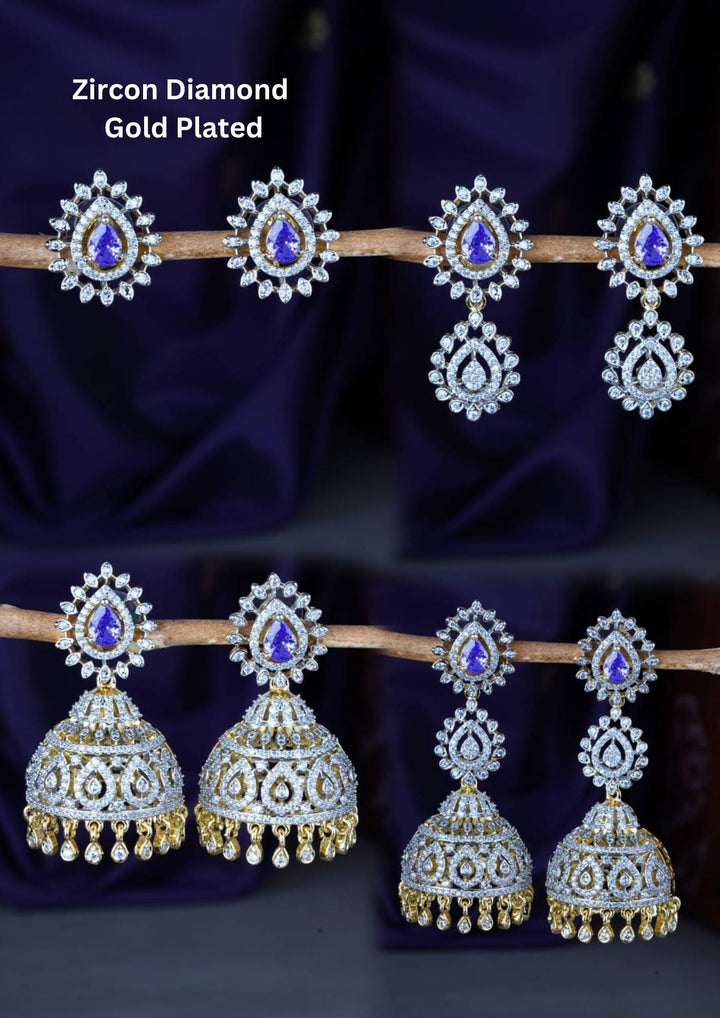 A premium collection of four distinct pairs of gold-plated earrings featuring brilliant white zircon diamonds and vibrant teardrop-shaped emerald blue stones. The assortment includes elegant halo stud earrings, multi-tiered crystal drops, and classic South Indian style hanging jhumkas adorned with intricate stone settings and tiny dangling gold beads, all beautifully arranged on a rustic branch against a deep purple fabric backdrop.