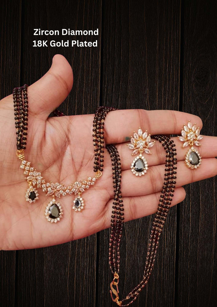 18K Gold Plated Zircon Diamond Mangalsutra set designed with a three-strand black bead Nallapusalu chain and a half moon-shaped pendant. The pendant show rows of sparkling white zircon stones in a radiating fringe pattern, paired with matching cluster stone stud earrings for a luxurious, premium jewelry finish. Perfect for any ocaasions and dailywaer