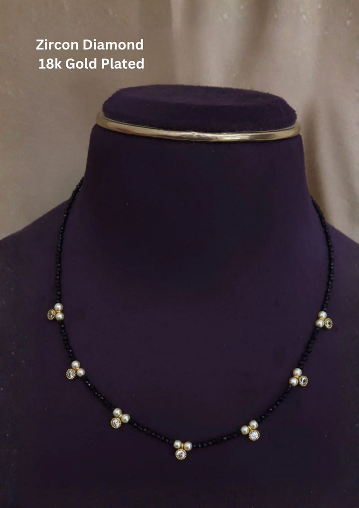 gold plated single-strand necklace composed of tiny, faceted black spinel beads. Spaced uniformly along the front section are seven delicate charm stations, each featuring a pair of small cream-white pearls flanking a central round-cut pure Zirconian diamond stone in a secure golden bezel.