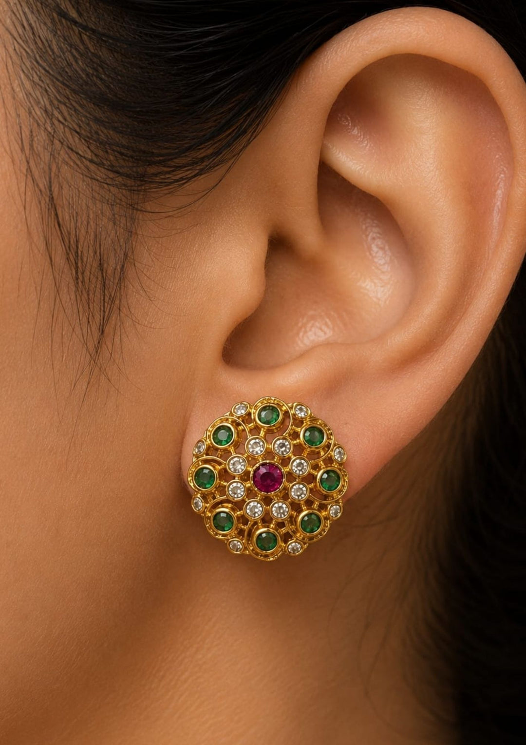 Close-up of an ear wearing a Traditional jadau kundan gold plated stud earrings featuring an intricate circular floral design crafted with radiant emerald-colored stones, sparkling kundan stones and a striking pink center stone. The symmetrical pattern and antique-inspired craftsmanship create a rich ethnic appeal suitable for weddings, festivals, poojas and traditional celebrations.