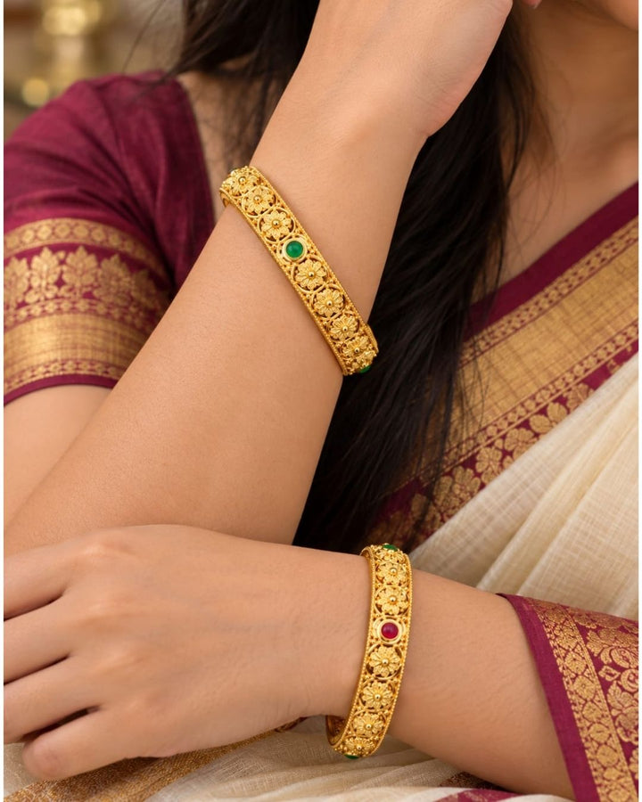 Elegant 18K gold plated floral bangles displayed on a carved wooden jewellery box, showcasing ornate gold craftsmanship and green and ruby stone details.