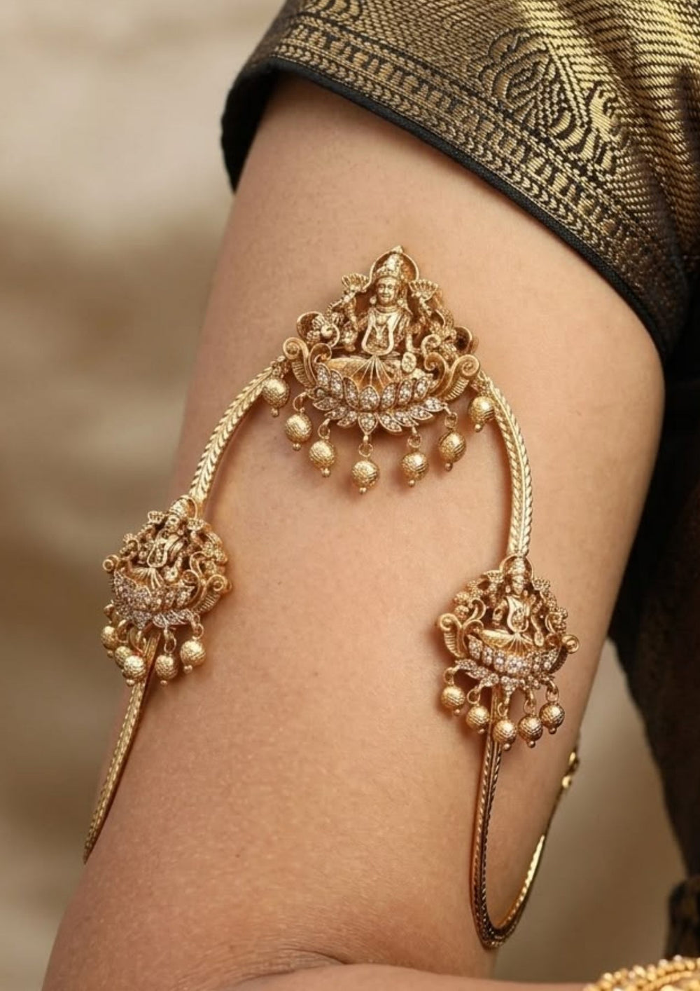 Gold plated vanki with intricate designs hand  A pair of traditional gold plated temple vanki armlets in image_990ae6.png featuring an elegant V-shaped braided band. Each armlet is adorned with central and side medallions embossed with Goddess Lakshmi seated on a lotus, accented with sparkling white crystals and dangling gold bead drops, displayed against a dark brown fabric background.