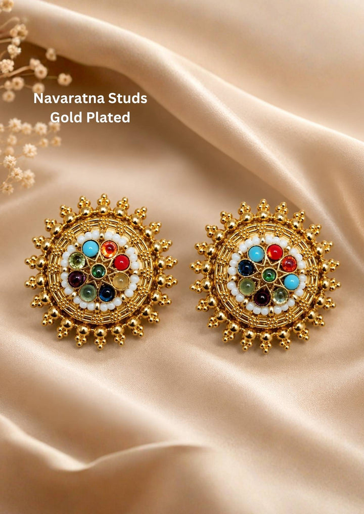 gold plated navaratna style round stud earrings featuring a wheel pattern arrangement of vibrant multi-color stones including turquoise blue, ruby red, emerald green, amethyst purple, and amber yellow bordered by fine white beads and an outer sunburst gold bead trim. These lightweight large statement studs provide a grand traditional look suitable for weddings, festivals, poojas, cultural events, festive sarees, lehengas, and ethnic outfits with skin-friendly quality, anti-fade polish,