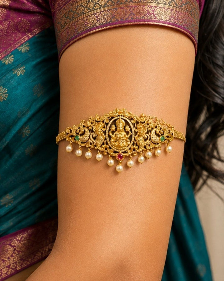 Premium gold plated 2-in-1 choker and bajuband featuring Goddess Lakshmi surrounded by intricate peacock and floral-inspired detailing, accented with green and pink stones and elegant pearl drops. Perfect for weddings, festive occasions, traditional wear, dance performances, parties and gifting.