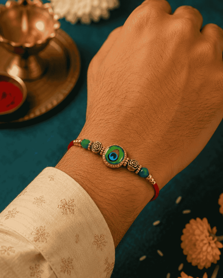 Traditional Bhaiya Bhabhi Rakhi set showcasing matching peacock-inspired charms, floral metal accents, green crystal beads, and an elegant dangling leaf charm.