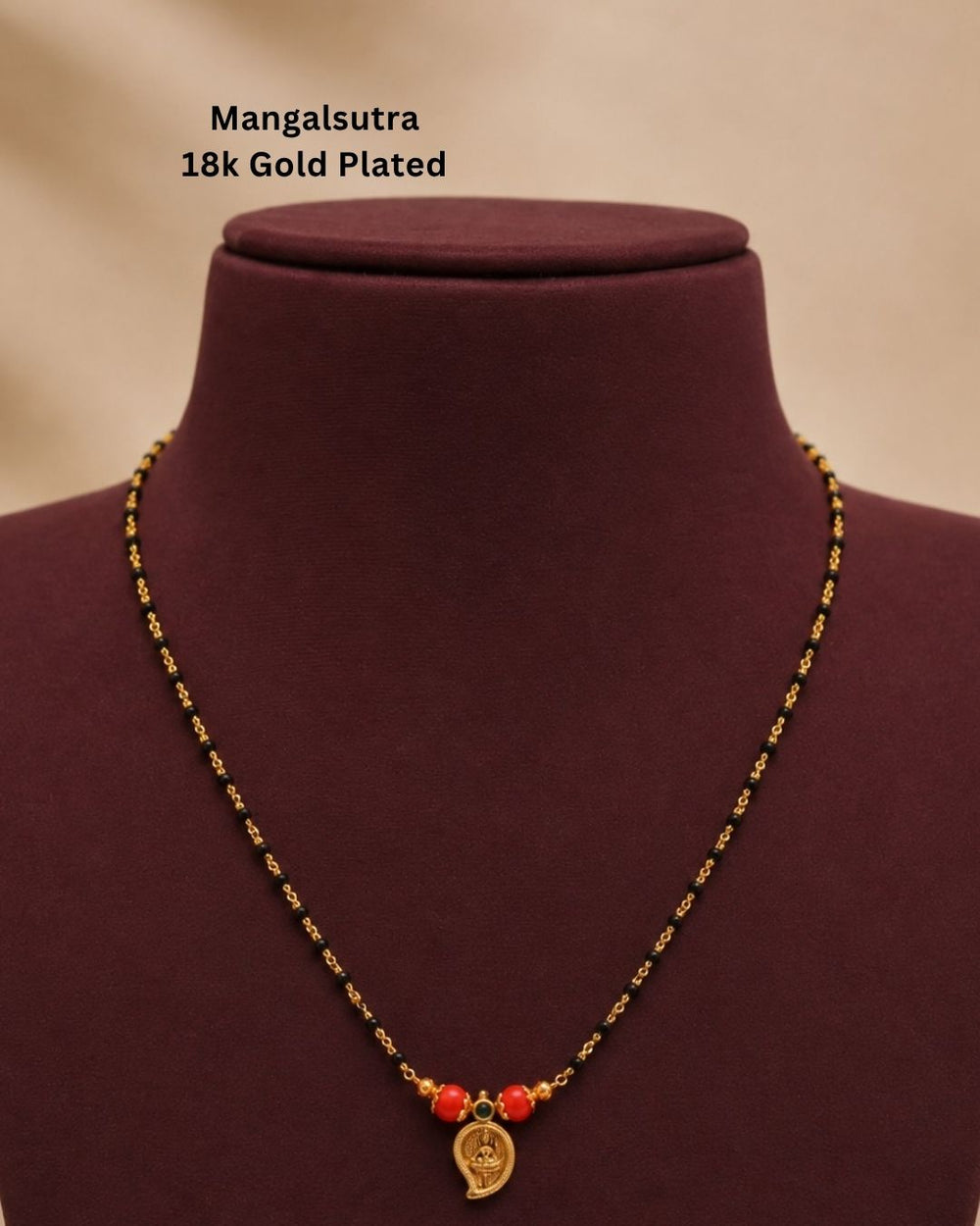 A gold-plated Mangalsutra necklace with red beads and a small gold pendant, displayed on a dark burgundy jewelry bust. Text in the upper left reads 'Mangalsutra 18k Gold Plated'.