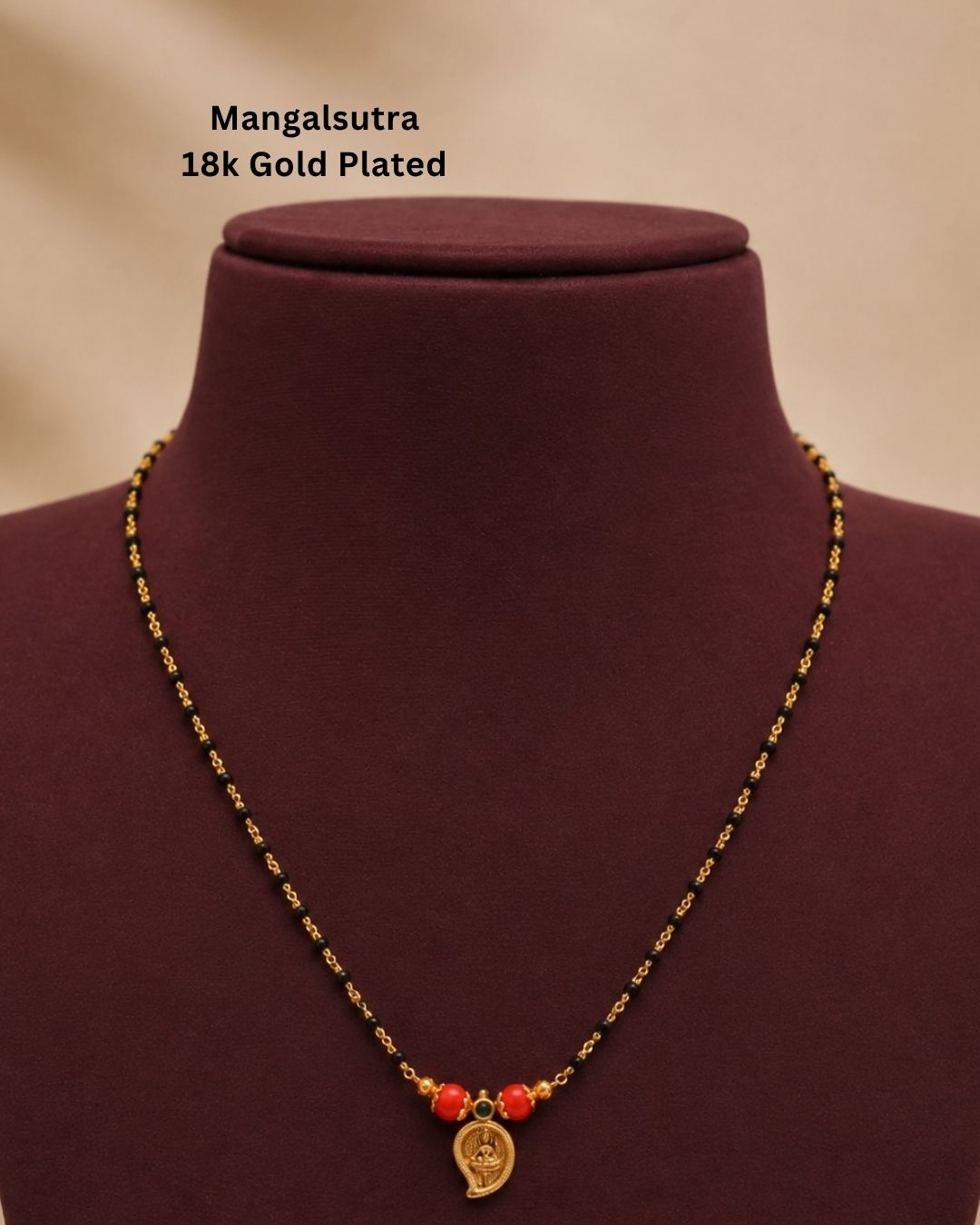 A gold-plated Mangalsutra necklace with red beads and a small gold pendant, displayed on a dark burgundy jewelry bust. Text in the upper left reads &