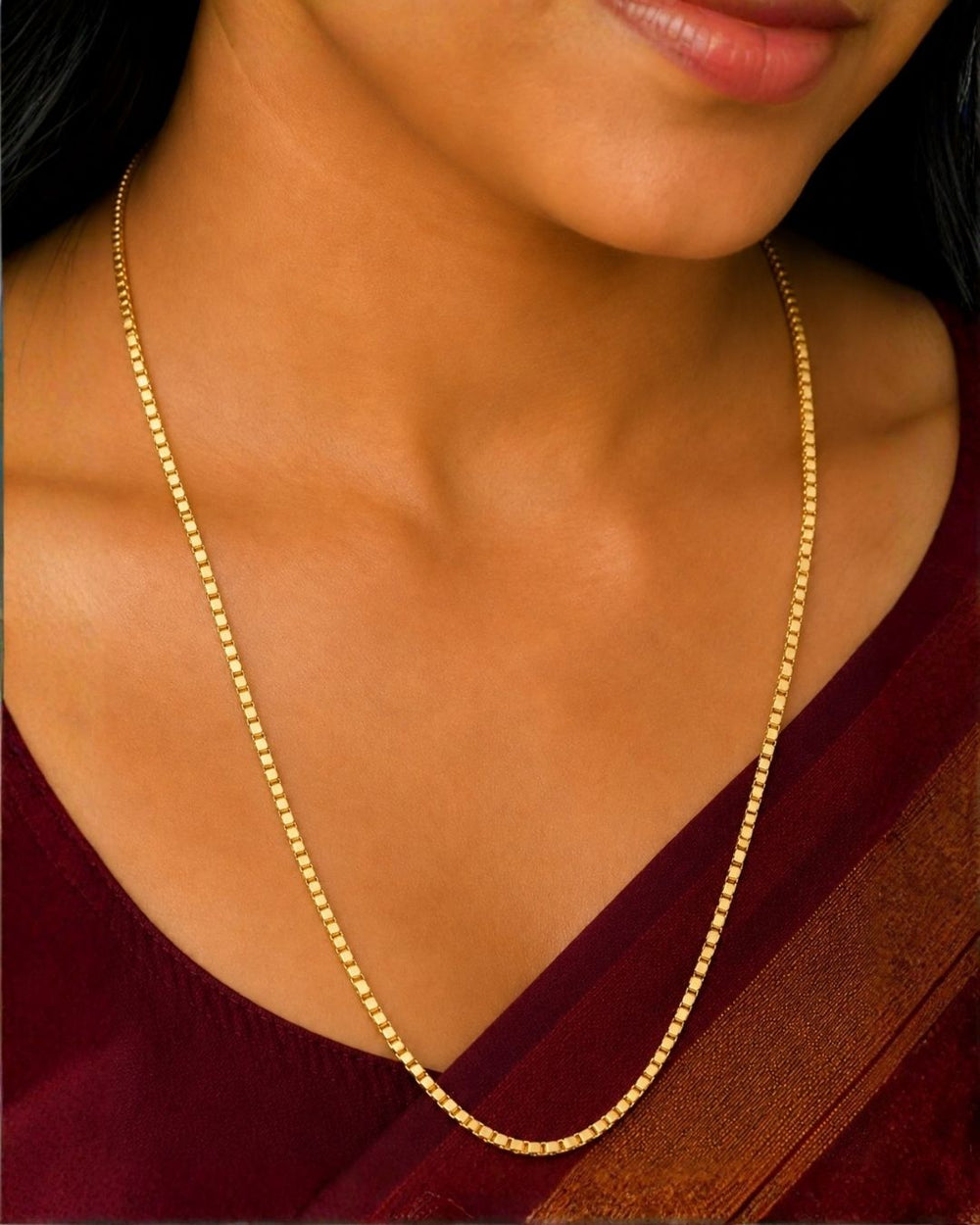 A close-up of a woman wearing a delicate gold chain necklace with small, tightly spaced beads. The necklace is displayed against her neck and upper chest, with a deep maroon sari visible behind it.