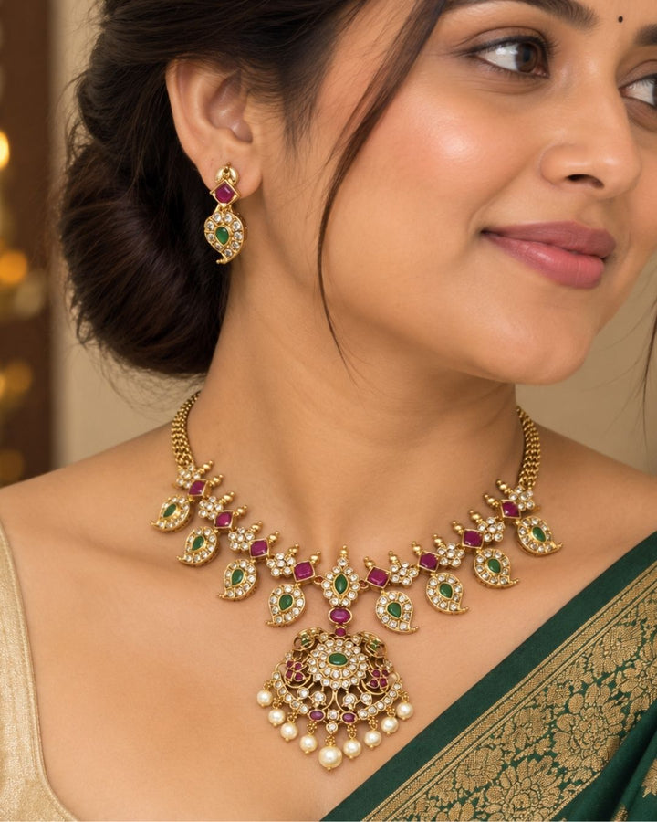 Traditional brass base 18K gold plated necklace set featuring detailed gold motifs, emerald and ruby stones, zircon embellishments, a statement pendant, pearl drops, and matching earrings.