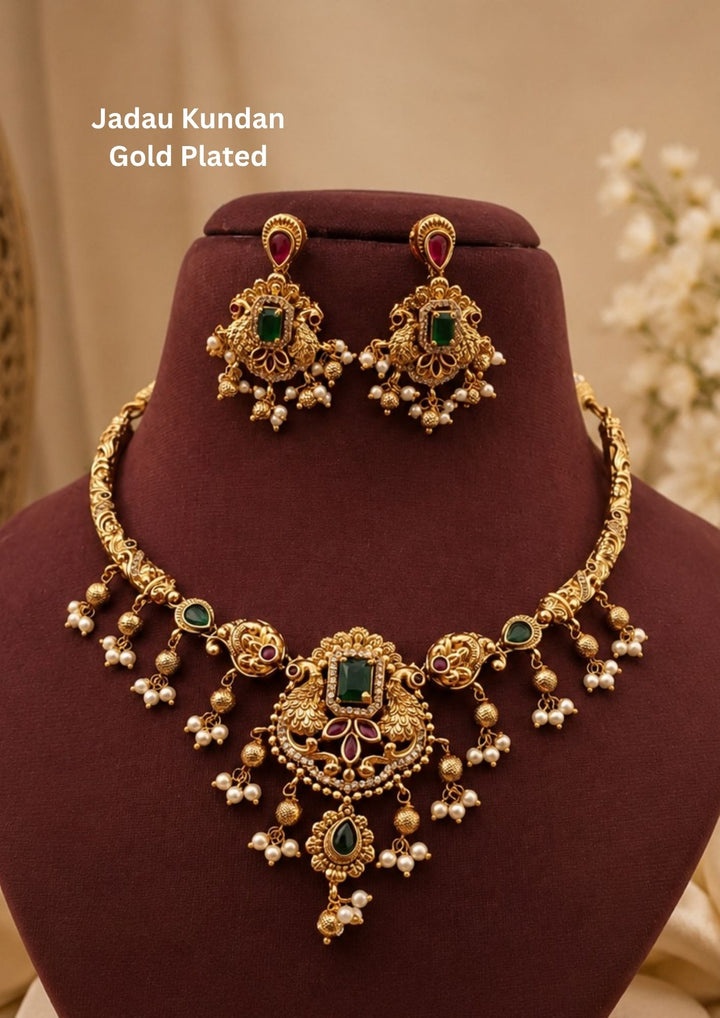 An exquisite Jadau Kundan gold plated necklace features a rigid gold-engraved band transitions into twin peacock motifs framing a central rectangular green emerald stone, accented by delicate hanging pearl clusters and matching drop earrings.