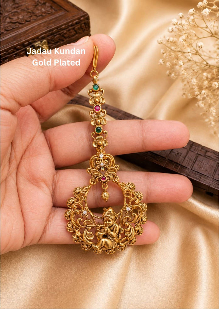 Gold plated traditional maang tikka hair accessory featuring a center medallion with a detailed, raised image of Lord Krishna playing the flute under a decorative archway. The pendant is outlined with sparkling white Kundan stones and finished with a neat row of small hanging white pearls at the bottom, providing a classic heritage look suitable for festive celebrations and family functions.