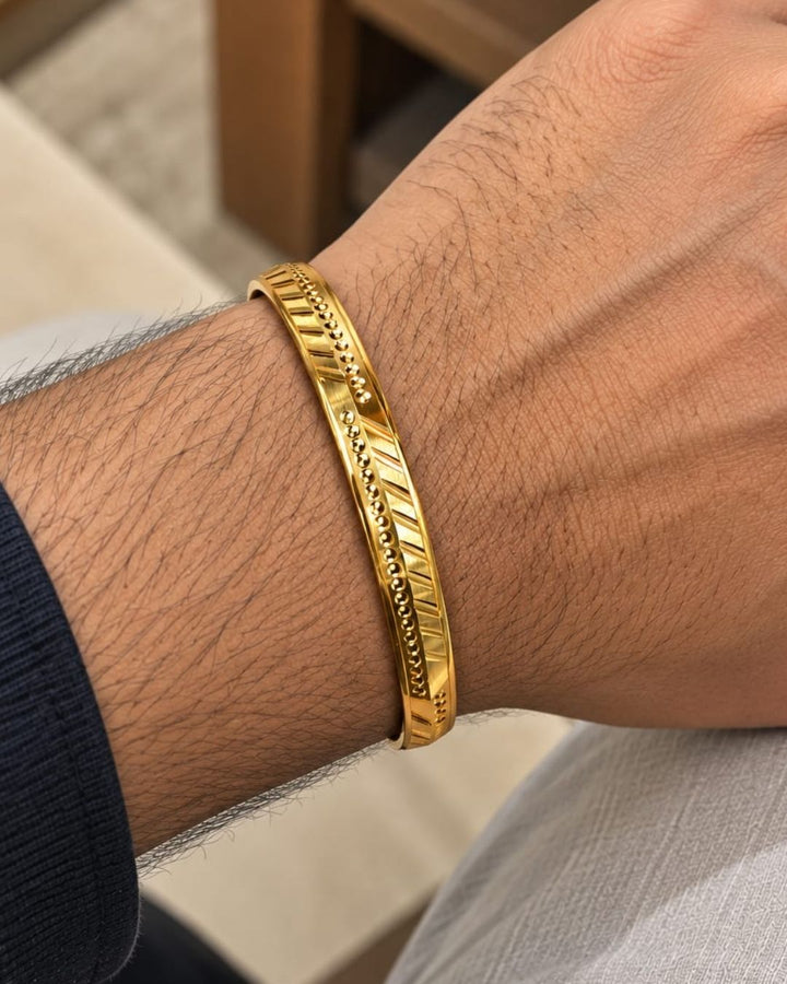 Gold plated men’s kada displayed upright on a dark textured surface, highlighting its geometric detailing and glossy finish.