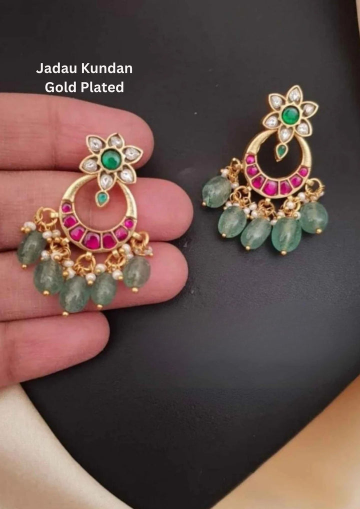 A pair of antique-style 18k gold plated chandbali earrings featuring a top flower-shaped stud with an emerald green core and white kundan petals. Hanging below is a hollow crescent moon framework inlaid with square ruby pink stones, finished with a fringe of tumbled green quartz beads and seed pearls.