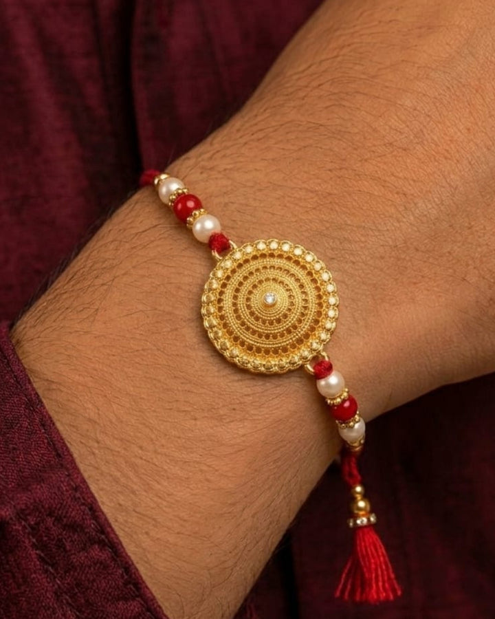 Elegant gold finish Rakhi showcasing a detailed circular mandala centerpiece with pearl and red bead embellishments on a vibrant red thread.