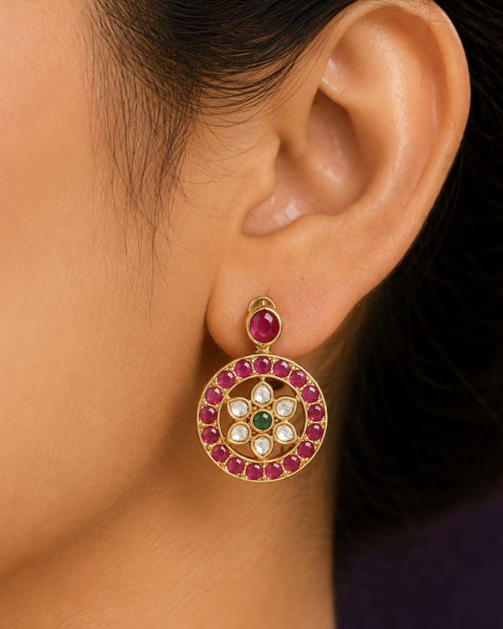 A close-up of a woman wearing a gold necklace and matching drop earrings with pink, green, and white floral details, paired with a dark purple sari.
