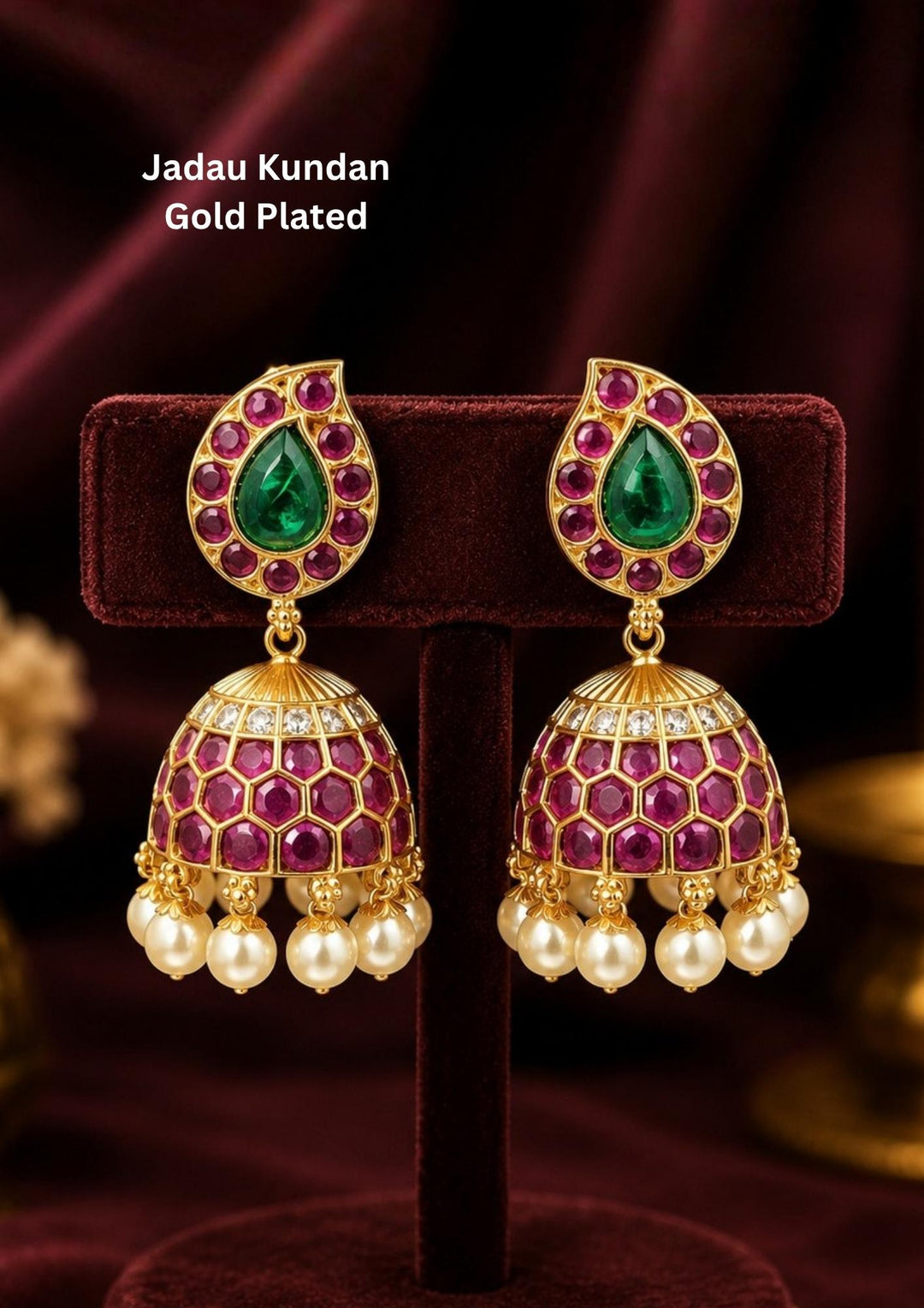Exquisite Jadau Kundan gold-plated jhumka earrings featuring a paisley or teardrop-shaped stud top. The top is centered with a striking green emerald stone framed by vibrant ruby-pink stones, leading down to a honeycomb-patterned dome adorned with pink stones and finished with elegant hanging pearl drops. Perfect for weddings and festive celebrations.