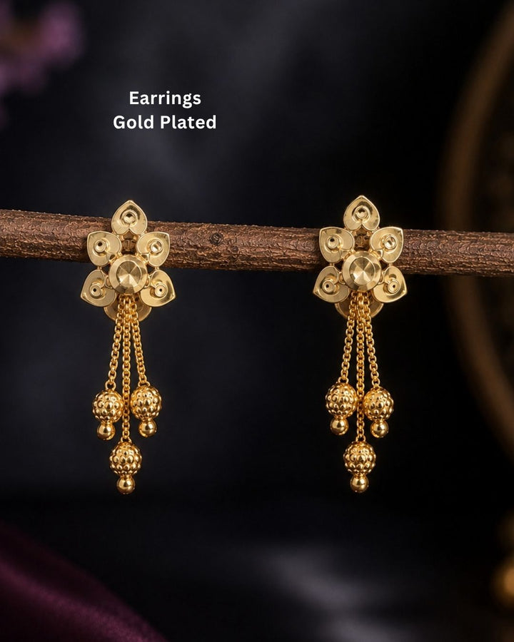 A pair of gold-plated traditional Indian jhumka earrings hanging from a wooden branch against a dark, blurred background. White text in the upper left reads &