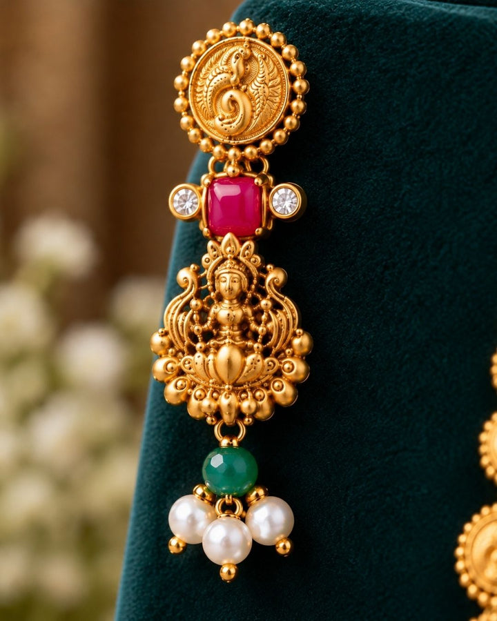 Elegant gold plated Lakshmi coin haram with detailed temple motifs, green beads, pearl drops, and matching Lakshmi earrings displayed on a teal jewellery bust.
