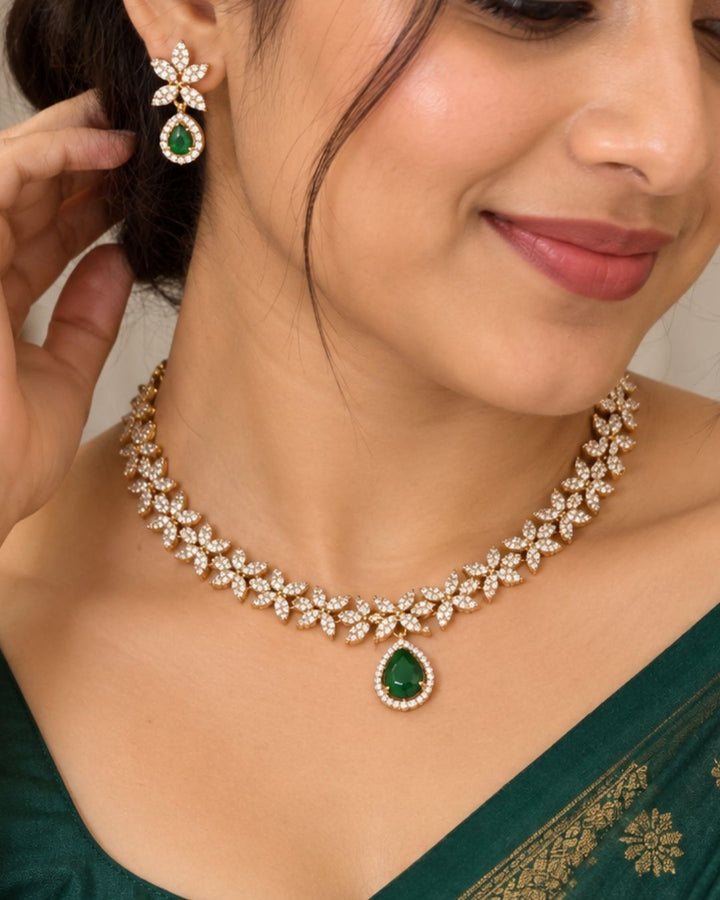 Designer gold plated necklace and earrings set featuring a delicate arrangement of leaf-shaped motifs embellished with sparkling white stones, a prominent green teardrop pendant, and matching green stone drop earrings, perfect for festive, wedding, and elegant occasions.