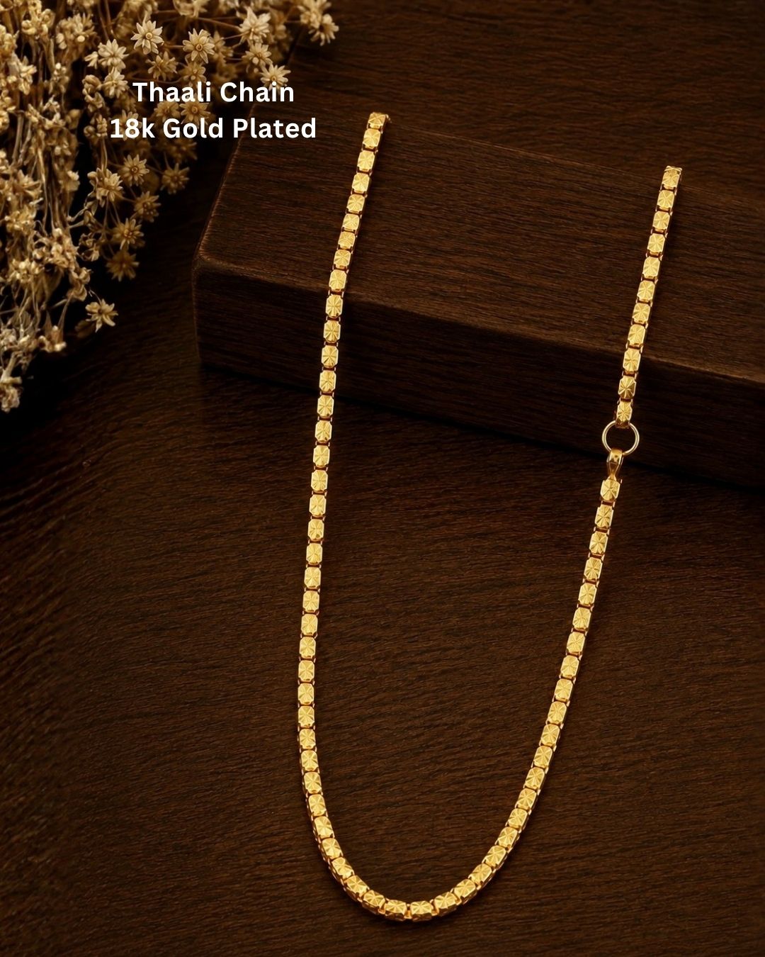 A 18K gold-plated Thaali chain necklace draped over a dark wooden block on a dark wood surface, with dried flowers in the top-left corner and text reading 'Thaali Chain 18K Gold Plated'.