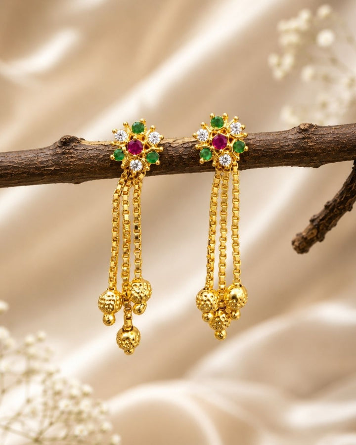 Stylish gold plated long drop earrings featuring vibrant ruby and green stones, sparkling white accents, intricate gold detailing, multiple hanging chains, and decorative gold ball drops. Perfect for festive occasions, traditional outfits, weddings, parties, and special events.