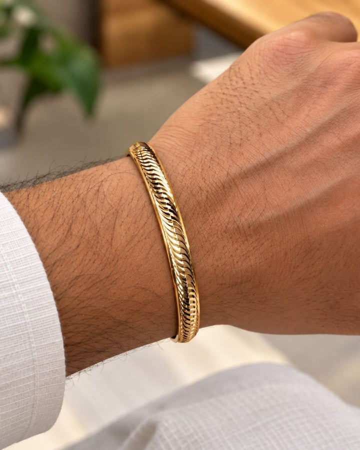 Gold plated men’s kada displayed upright on a beige surface with a soft pink background, highlighting its woven wave design and polished finish.