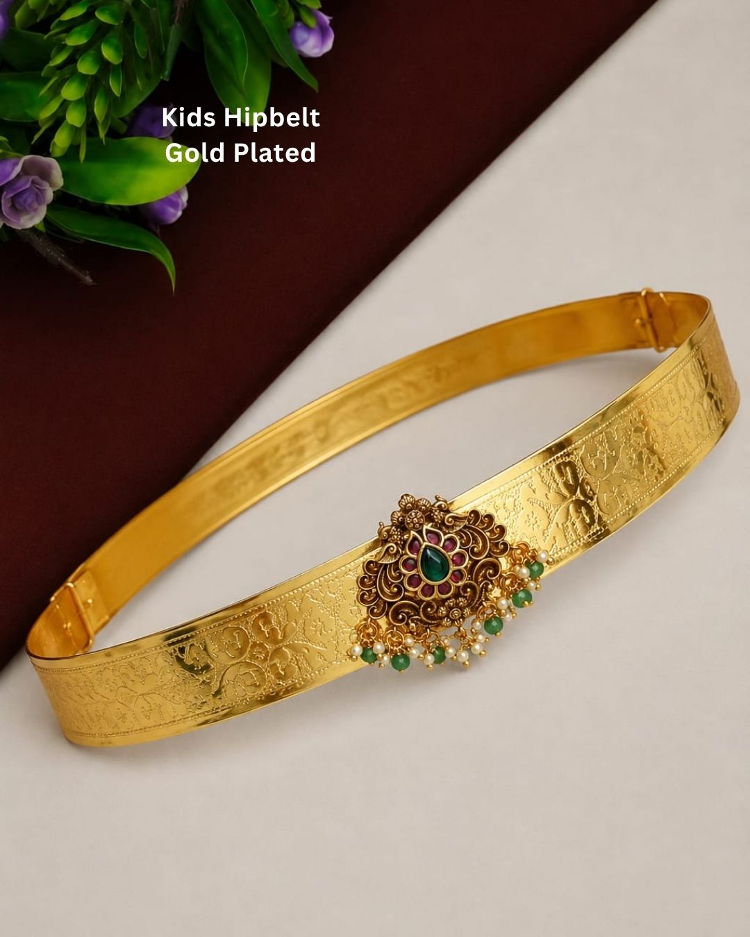 Beautiful gold plated kid’s hip belt featuring a wide intricately engraved band with a floral gemstone centerpiece decorated with ruby, green stones, and pearl drops. Perfect for children’s traditional wear, weddings, festive occasions, cultural events, parties, and gifting.