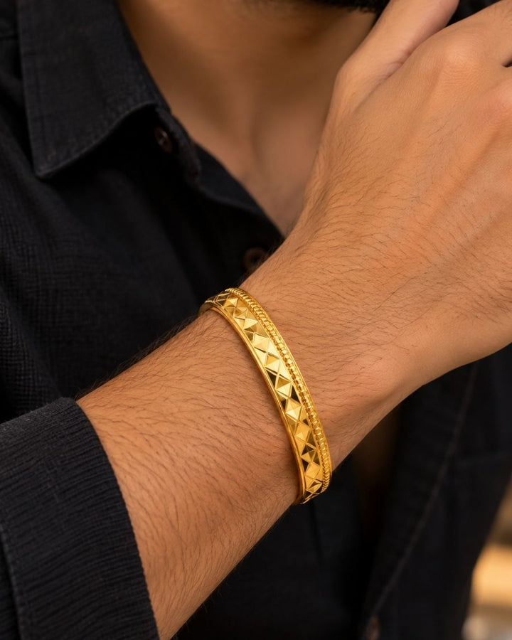 Gold plated men’s kada displayed upright on a warm beige surface, showcasing its diamond-cut design and glossy finish.