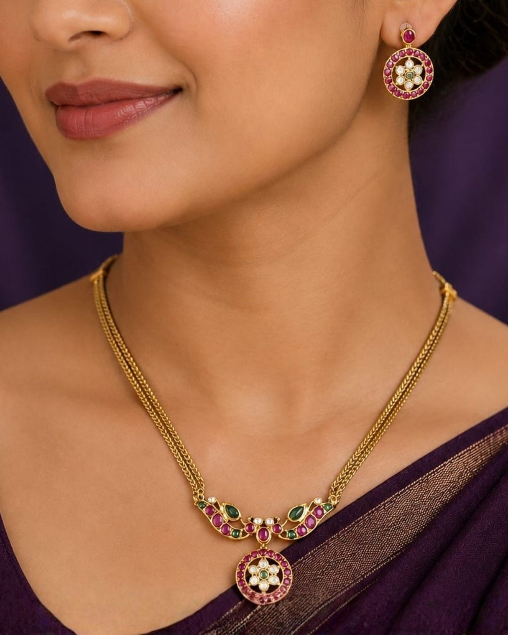 A close-up of a woman wearing a gold necklace and matching drop earrings with pink, green, and white floral details, paired with a dark purple sari.
