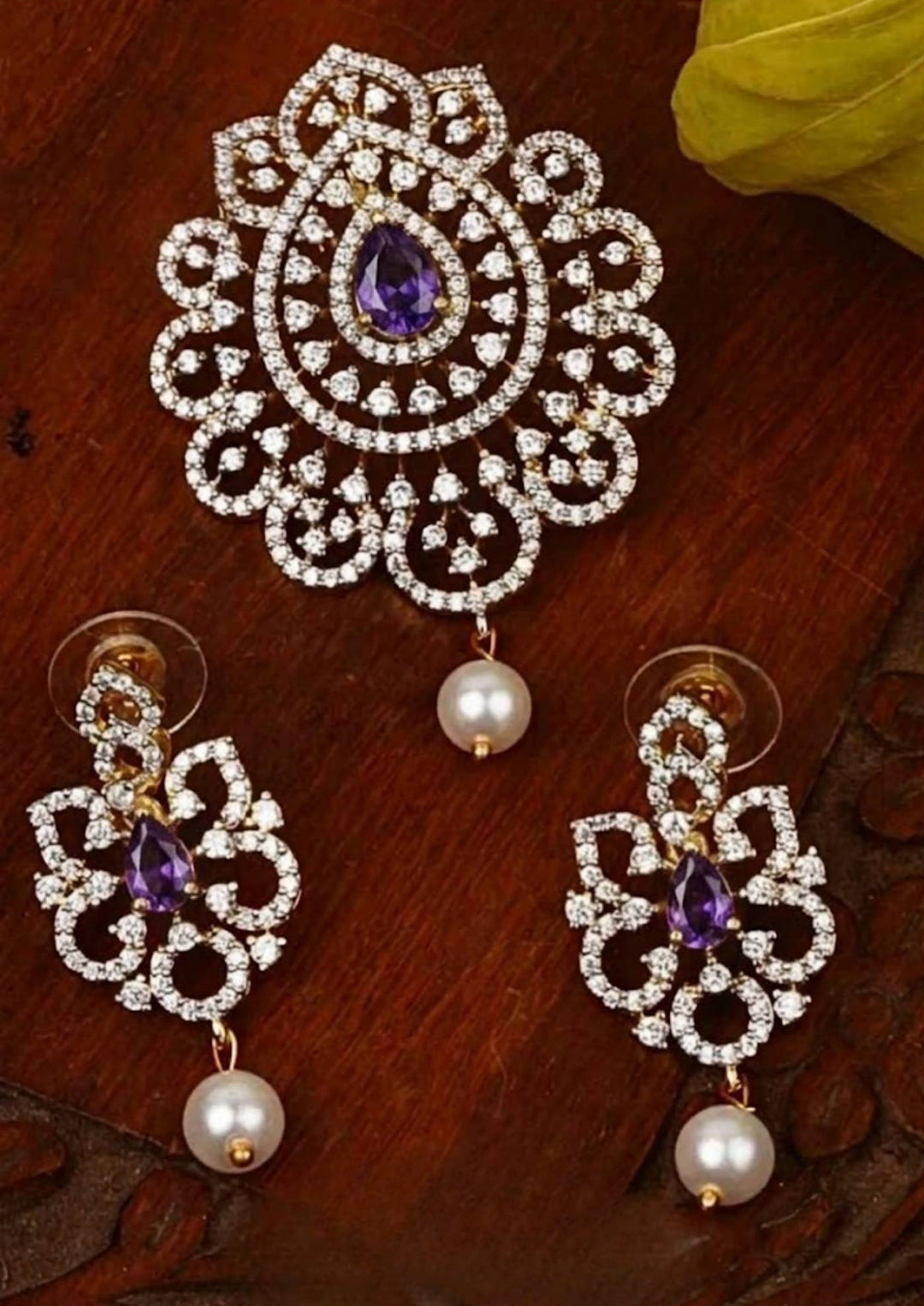 Elegant zircon diamond gold plated pendant set featuring a floral-inspired pendant designed with sparkling white zircon stones arranged in circular patterns. The centerpiece showcases a pear-shaped emerald violet stone surrounded by dazzling zircon stones, finished with a delicate pearl drop. The matching earrings mirror the same floral design with green stone and pearl danglers, Perfect for weddings, festivals, office wear, functions and its anti fade color