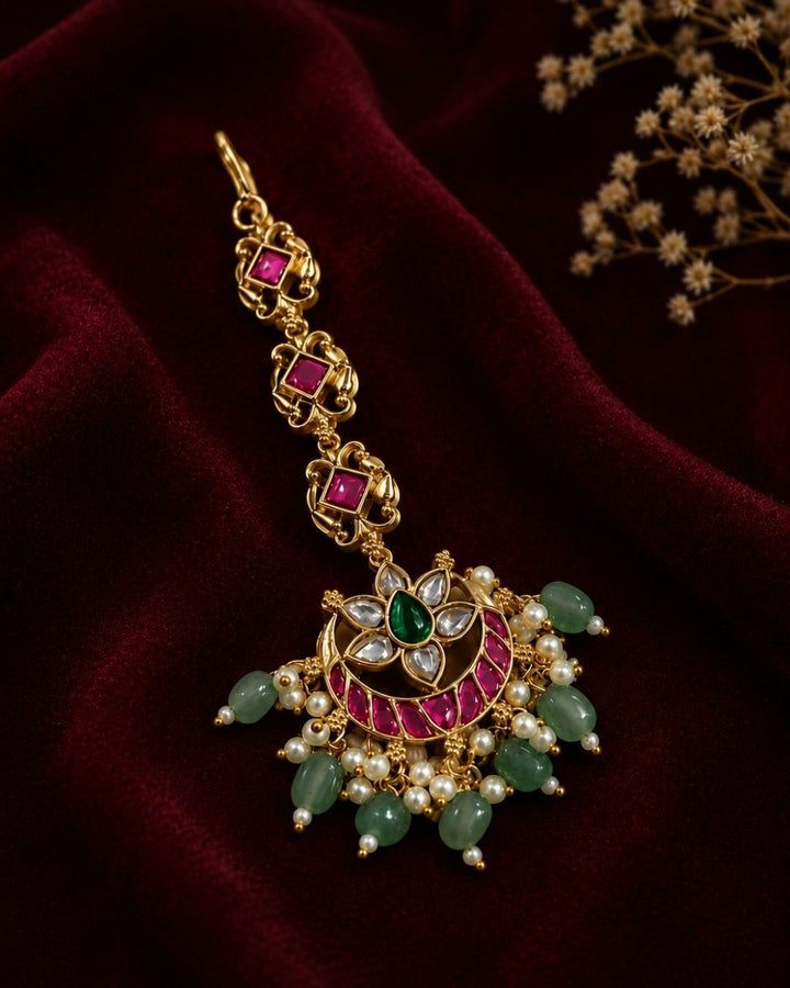 Beautiful traditional gold plated maang tikka featuring a pink stone chain, a floral-inspired circular pendant with green and pink stones, sparkling white accents, clusters of pearls, and multiple green bead drops. Perfect for weddings, festive occasions, traditional outfits, bridal styling, and special celebrations.