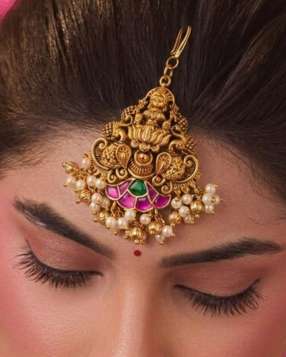An ornate gold-plated Maangtikka hair pendant with intricate filigree work, small pearl-like beads, and pink and green stone accents, displayed on a beige satin background with text reading 'Maangtikka 18k Gold Plated'.