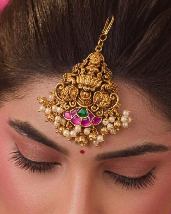 An ornate gold-plated Maangtikka hair pendant with intricate filigree work, small pearl-like beads, and pink and green stone accents, displayed on a beige satin background with text reading &