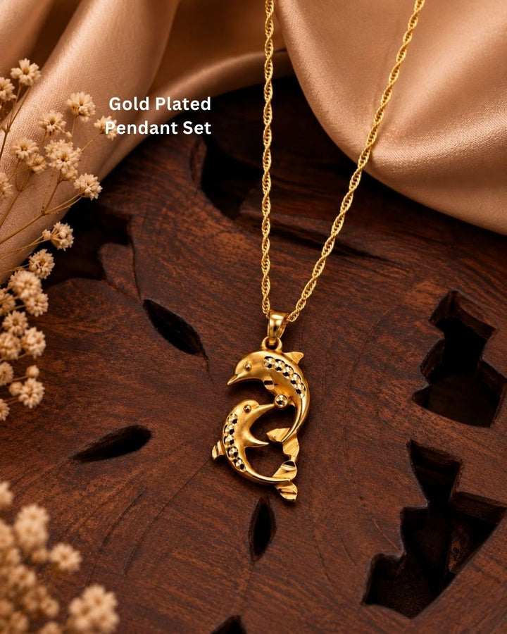 Gold-plated pendant necklace featuring a dolphin design with small clear stones, displayed on a dark wooden surface with beige satin fabric and dried flowers. Text overlay reads &