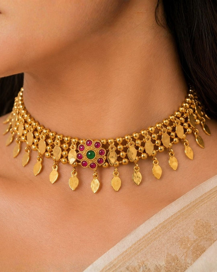 Gold-toned traditional Indian jewelry set featuring a choker necklace and matching drop earrings worn by a model. The jewelry is adorned with gold leaf-shaped charms, small beads, and central floral motifs set with green and red stones.
