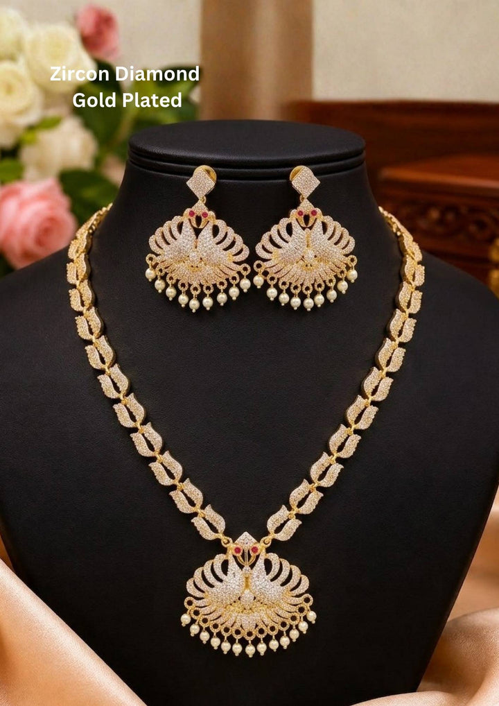 Premium gold plated jewelry set featuring a medium-length link chain decorated with fine white zircon stones and a grand twin peacock pendant arranged in a fan-like wing design. The centerpiece is highlighted with tiny ruby red stone eyes and a delicate fringe of hanging white pearls at the base. It comes complete with matching large peacock drop earrings, crafted with skin-friendly quality