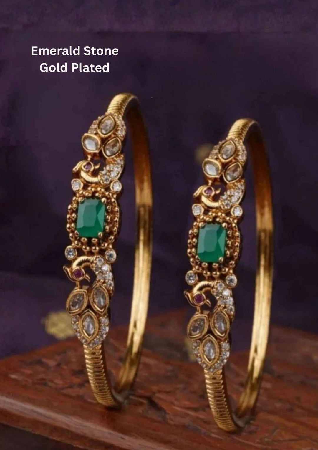A pair of premium 18k gold plated kada bangles featuring an exquisite twin peacock design flanking a central rectangular emerald green gemstone. The peacock silhouettes are delicately studded with tiny shimmering white stones and accented with ruby red eyes. The remaining portion of the band features a clean, highly polished ribbed texture.