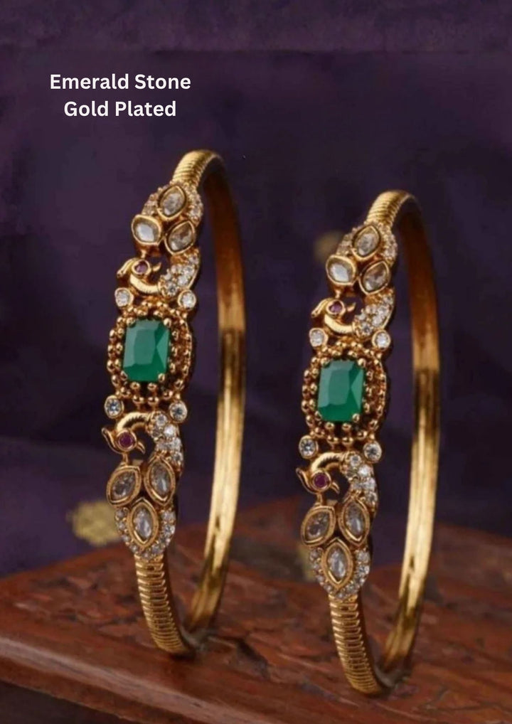 A pair of premium 18k gold plated kada bangles featuring an exquisite twin peacock design flanking a central rectangular emerald green gemstone. The peacock silhouettes are delicately studded with tiny shimmering white stones and accented with ruby red eyes. The remaining portion of the band features a clean, highly polished ribbed texture.