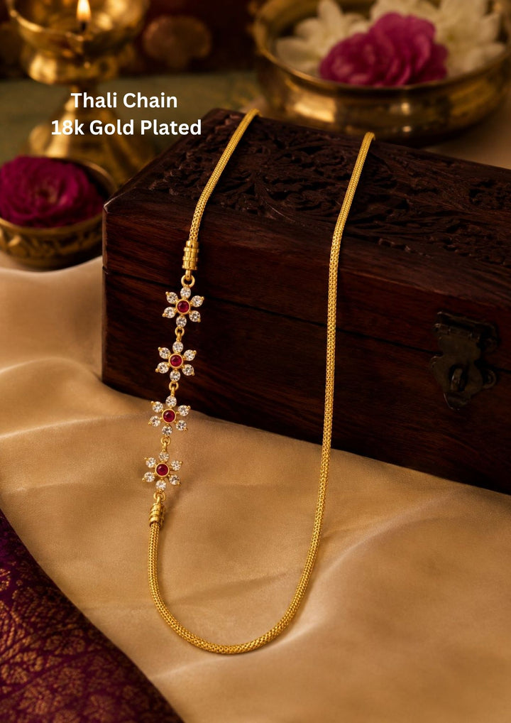 18k gold plated thali chain featuring a distinctive side mogappu pendant designed as a vertical row of four connected flowers, each meticulously crafted with sparkling white zircon petals and a deep ruby pink central stone. This delicate woven gold chain offers a premium, elongated boutique appearance suitable for weddings, festive celebrations, poojas, family milestone events, traditional sarees, and bridal wear with skin-friendly quality, anti-fade long-lasting polish, lightweight comfort