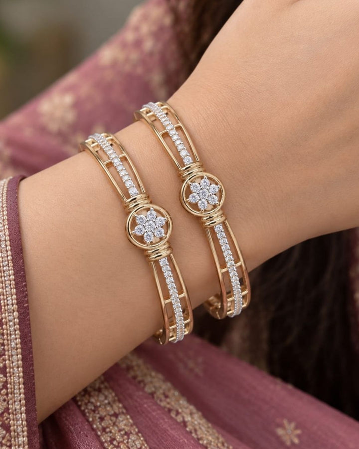 Pair of gold plated designer bangles displayed upright on a dark wooden surface with a rich brown background.