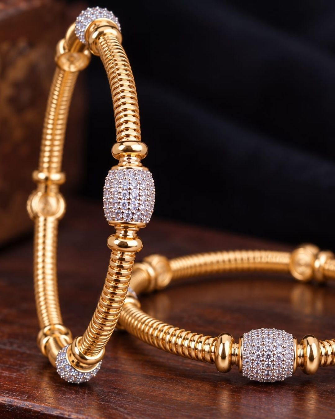 Pair of gold plated designer bangles displayed on a dark wooden surface against a black fabric background.