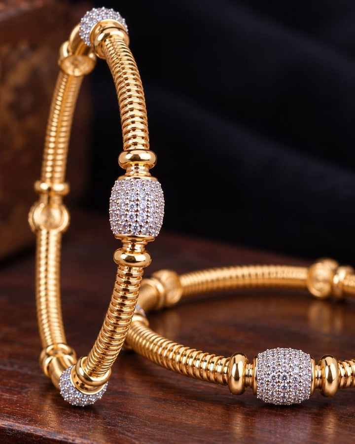 Pair of gold plated designer bangles displayed on a dark wooden surface against a black fabric background.
