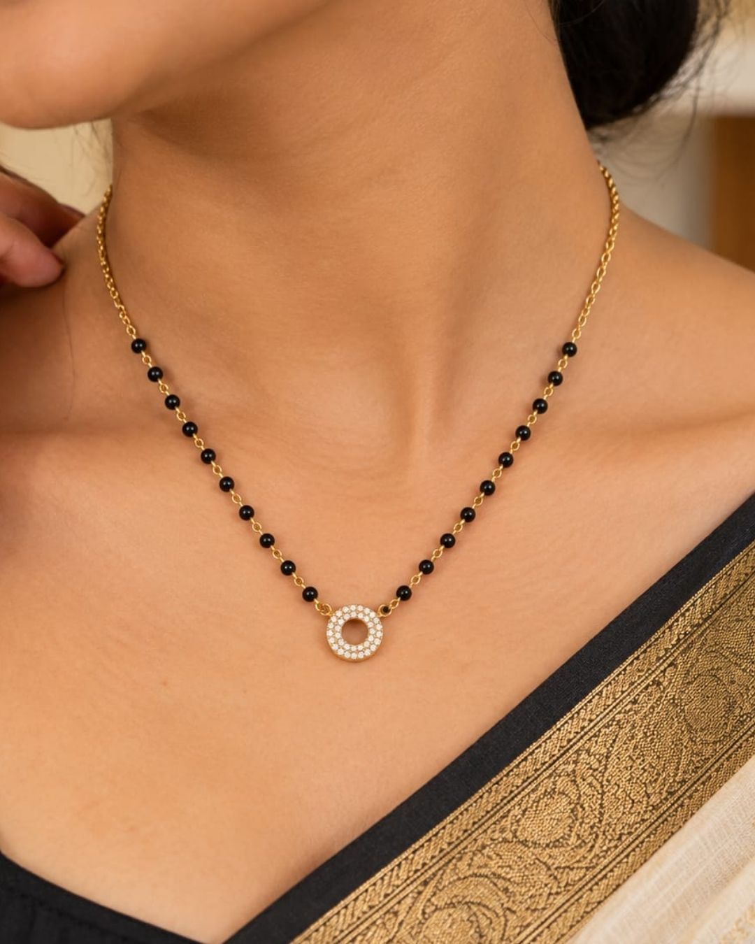 South indian Women wearing Gold plated black bead mangalsutra with a circular stone pendant displayed on a beige surface with a minimal decorative background.