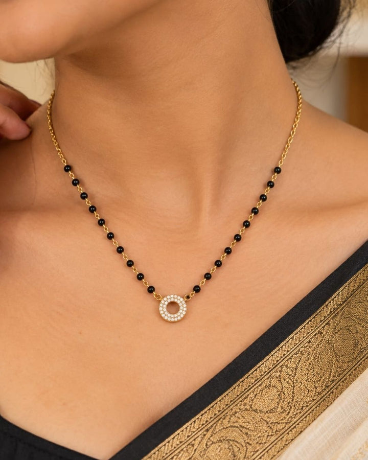 South indian Women wearing Gold plated black bead mangalsutra with a circular stone pendant displayed on a beige surface with a minimal decorative background.