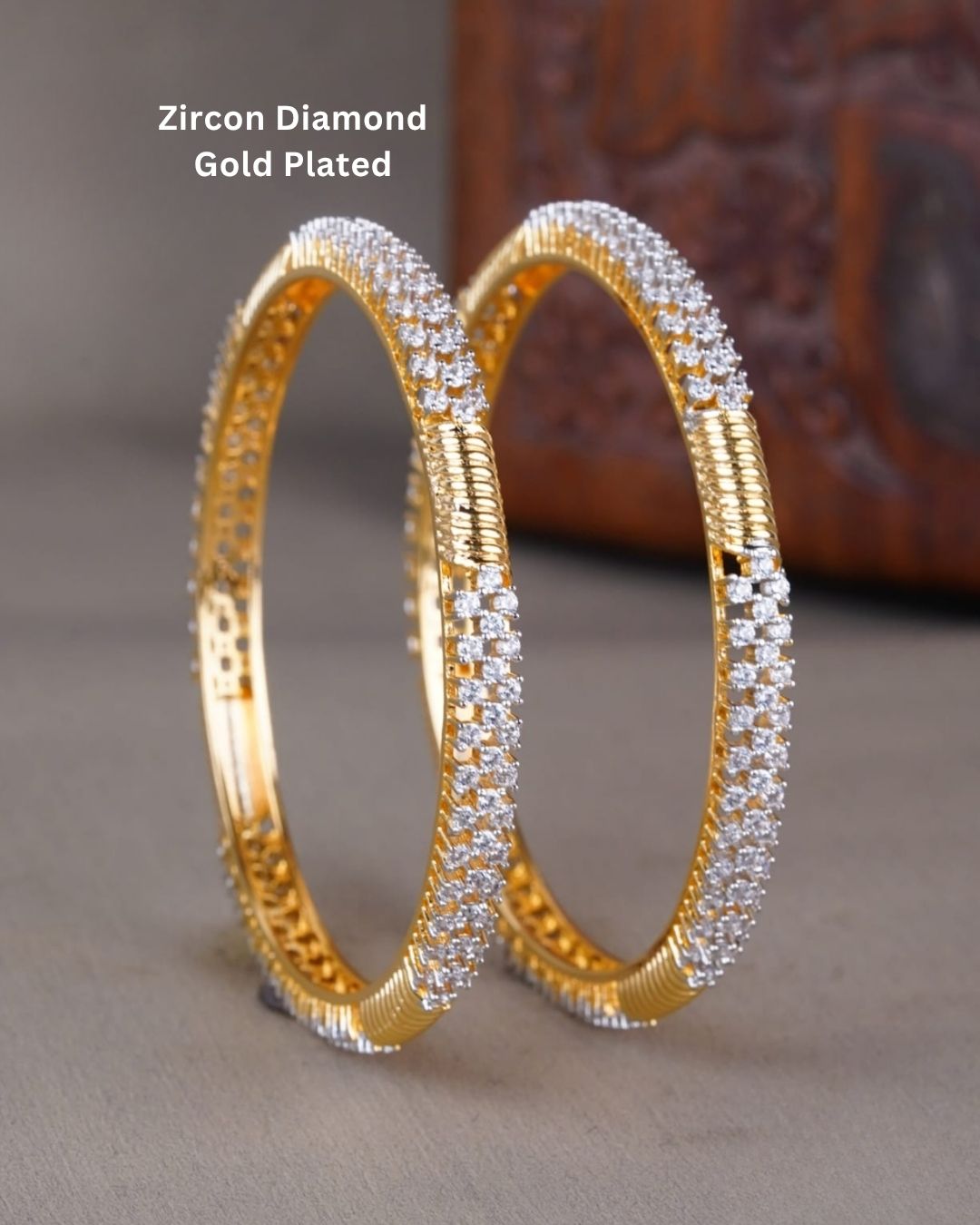 A pair of gold-plated huggie bangles adorned with rows of sparkling clear zirconia stones, displayed on a neutral surface with a blurred wooden background. Text overlay reads 'Zircon Diamond Gold Plated'.